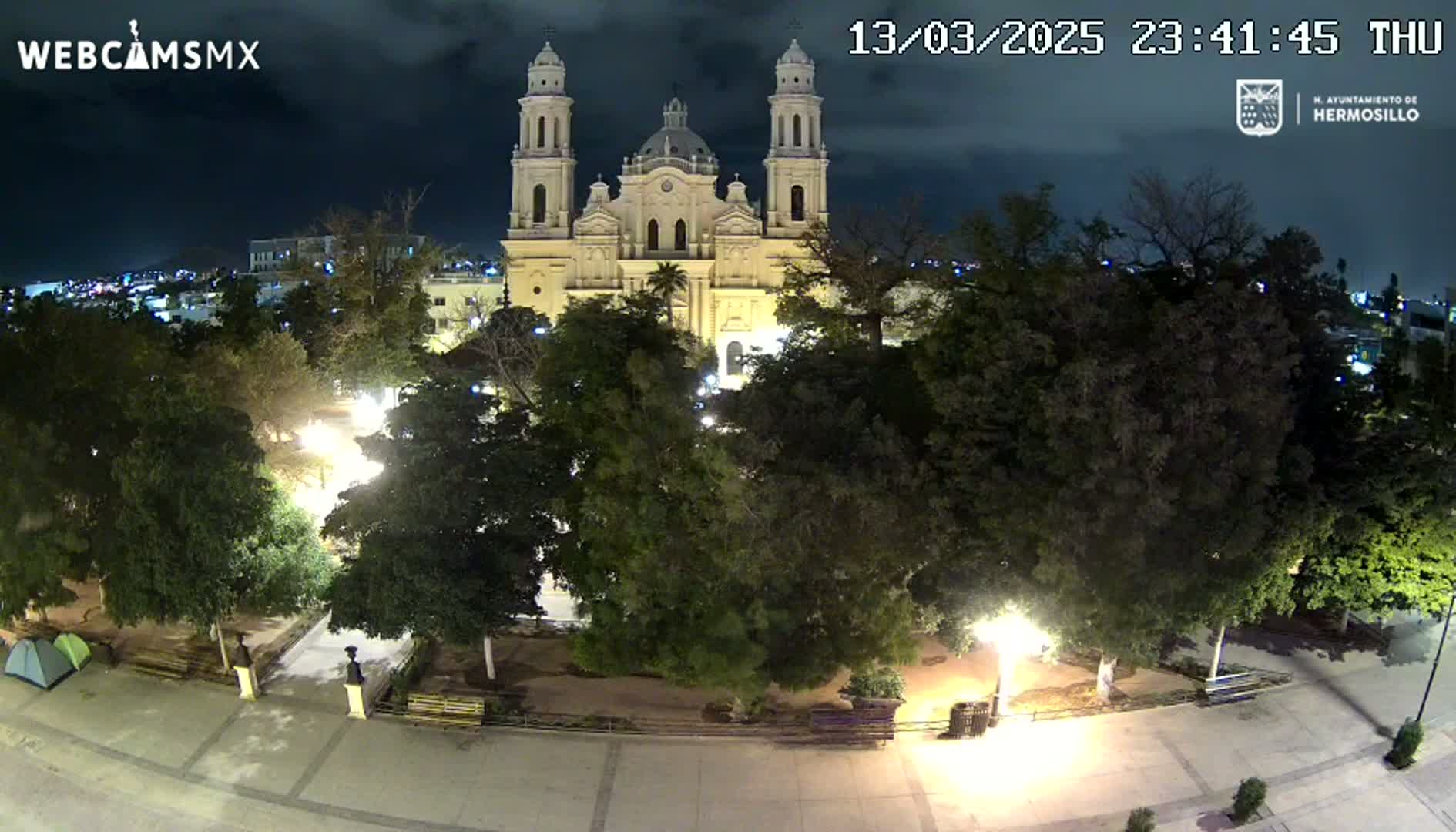 A nighttime, overhead view of a plaza with a large cathedral in the background under a partly cloudy sky.
