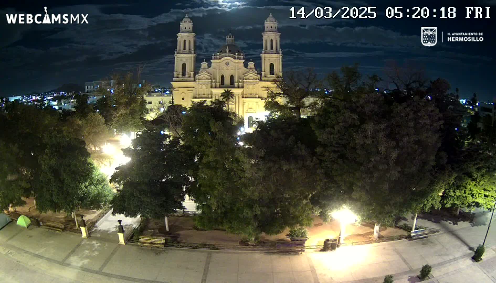 A nighttime, moonlit view of a town square with a large cathedral in the background, surrounded by lush green trees and illuminated by streetlights.