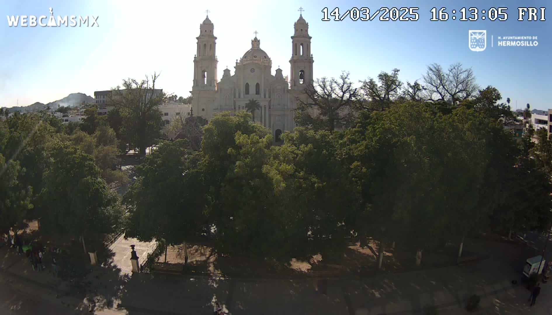 A large, light-colored cathedral stands behind a park filled with leafy green trees under a sunny sky.