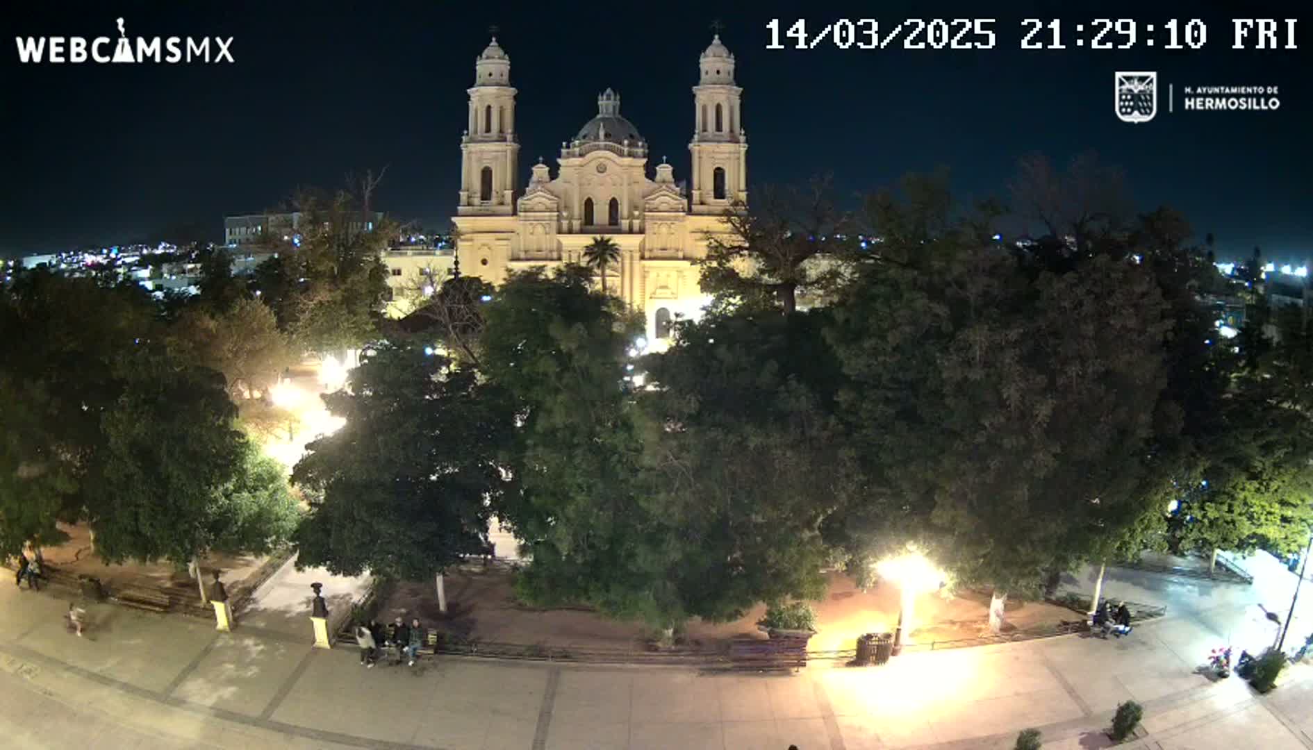 A nighttime view of a town square with a large church at its center, surrounded by lush green trees and illuminated by streetlights.