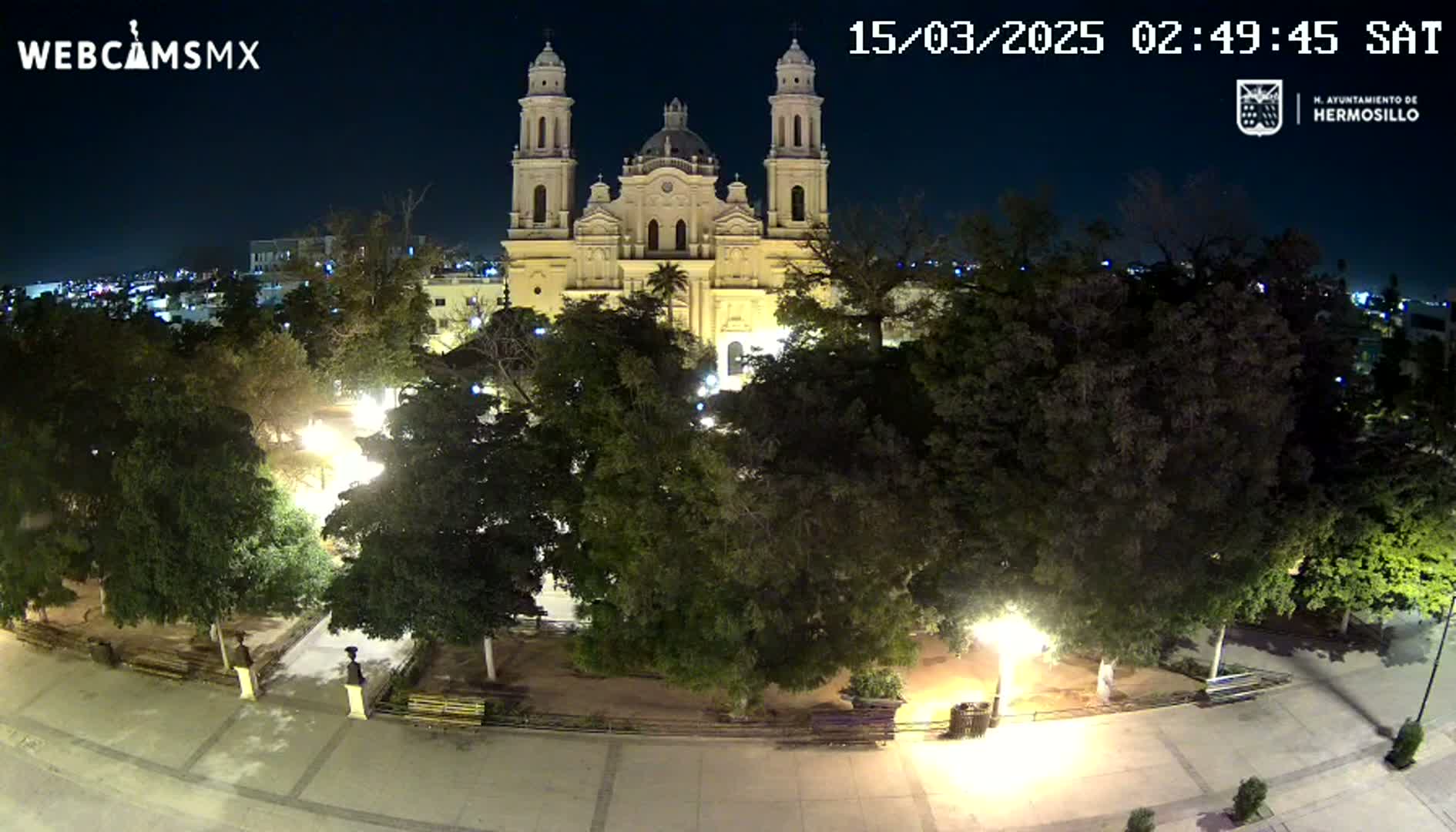 A brightly lit plaza at night features a large cathedral in the background, surrounded by trees and benches.