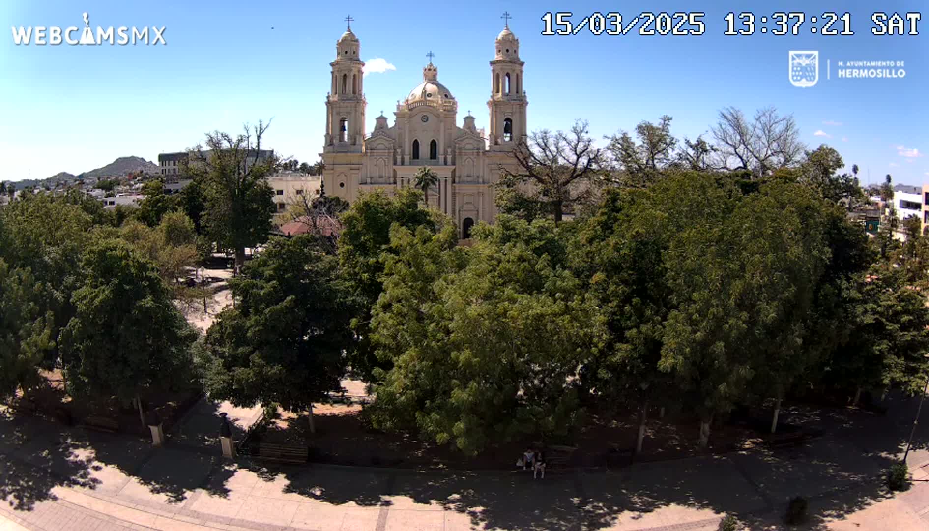 A large tan cathedral is visible behind a lush green park under a clear blue sky.