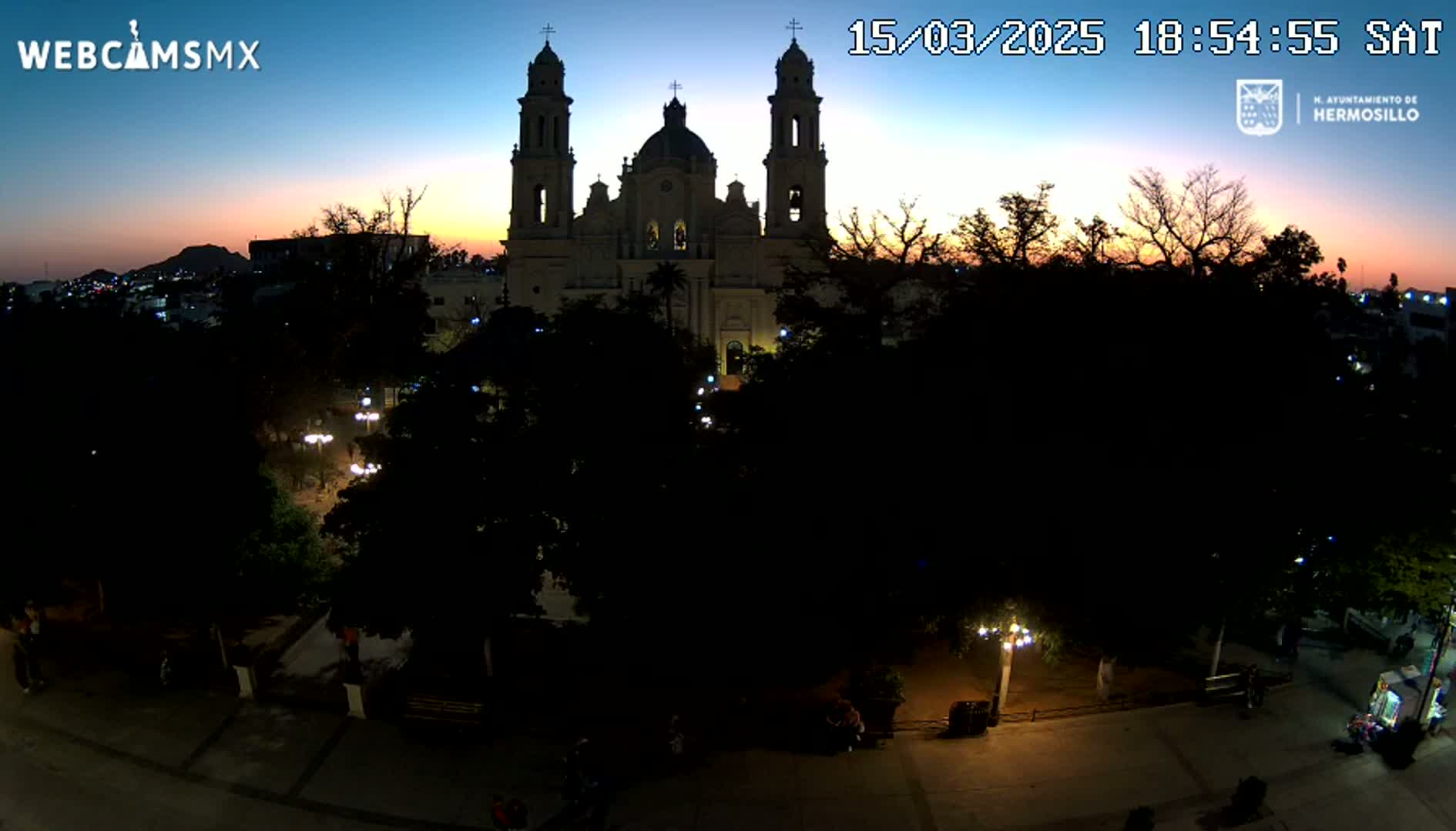 A large cathedral is silhouetted against a twilight sky, with a city square and surrounding trees visible below in the dimly lit night.