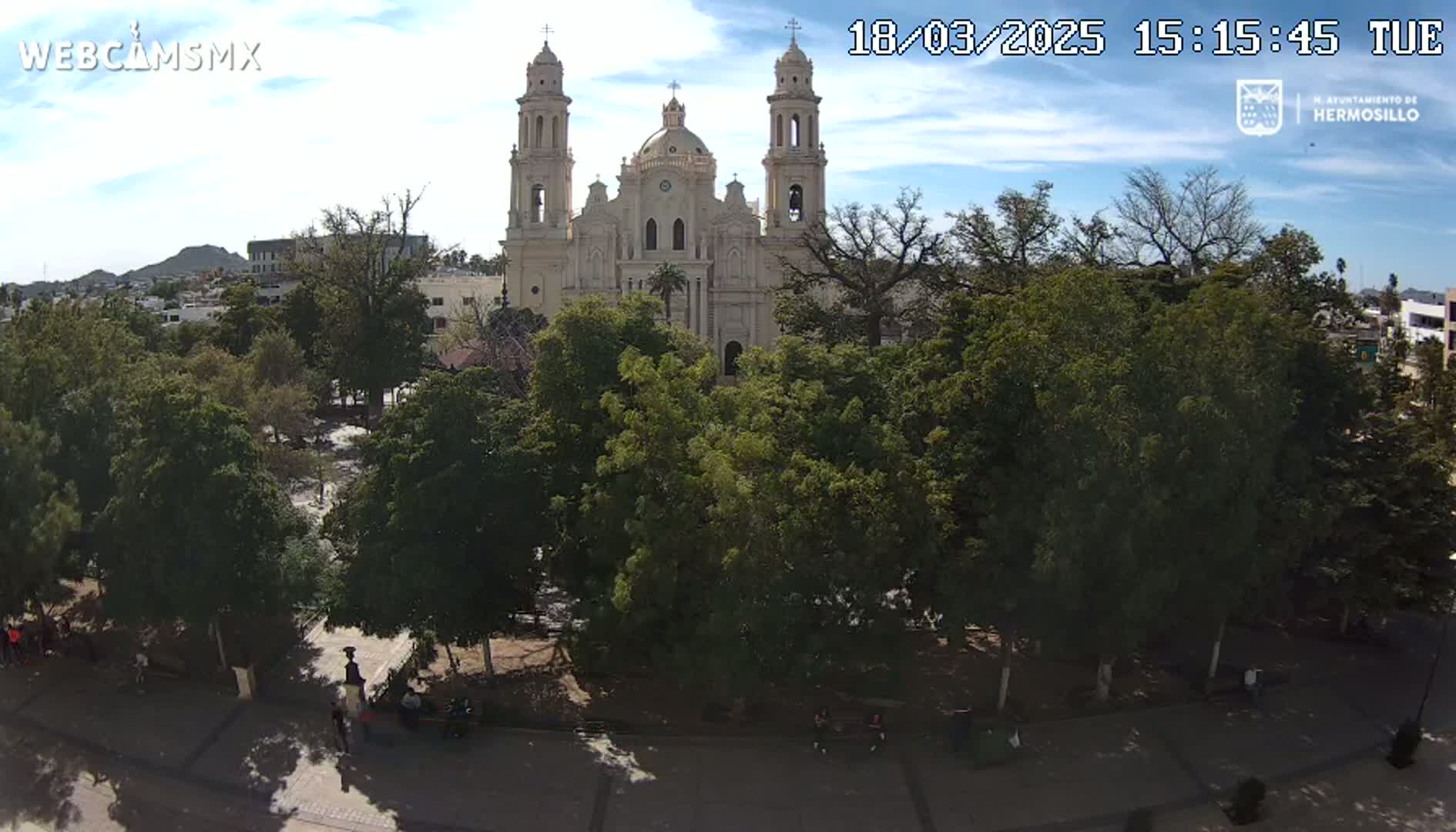 A large cathedral is visible behind a plaza filled with lush green trees under a partly cloudy sky.