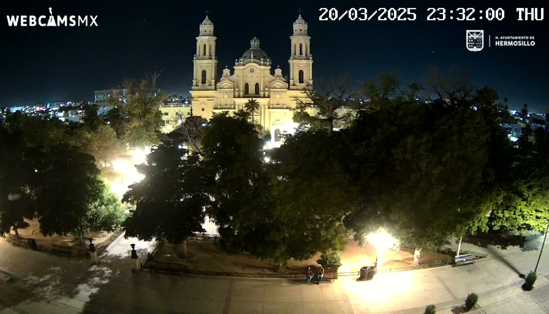 A brightly lit cathedral is visible at night, surrounded by a park with trees and pathways, under a clear sky.