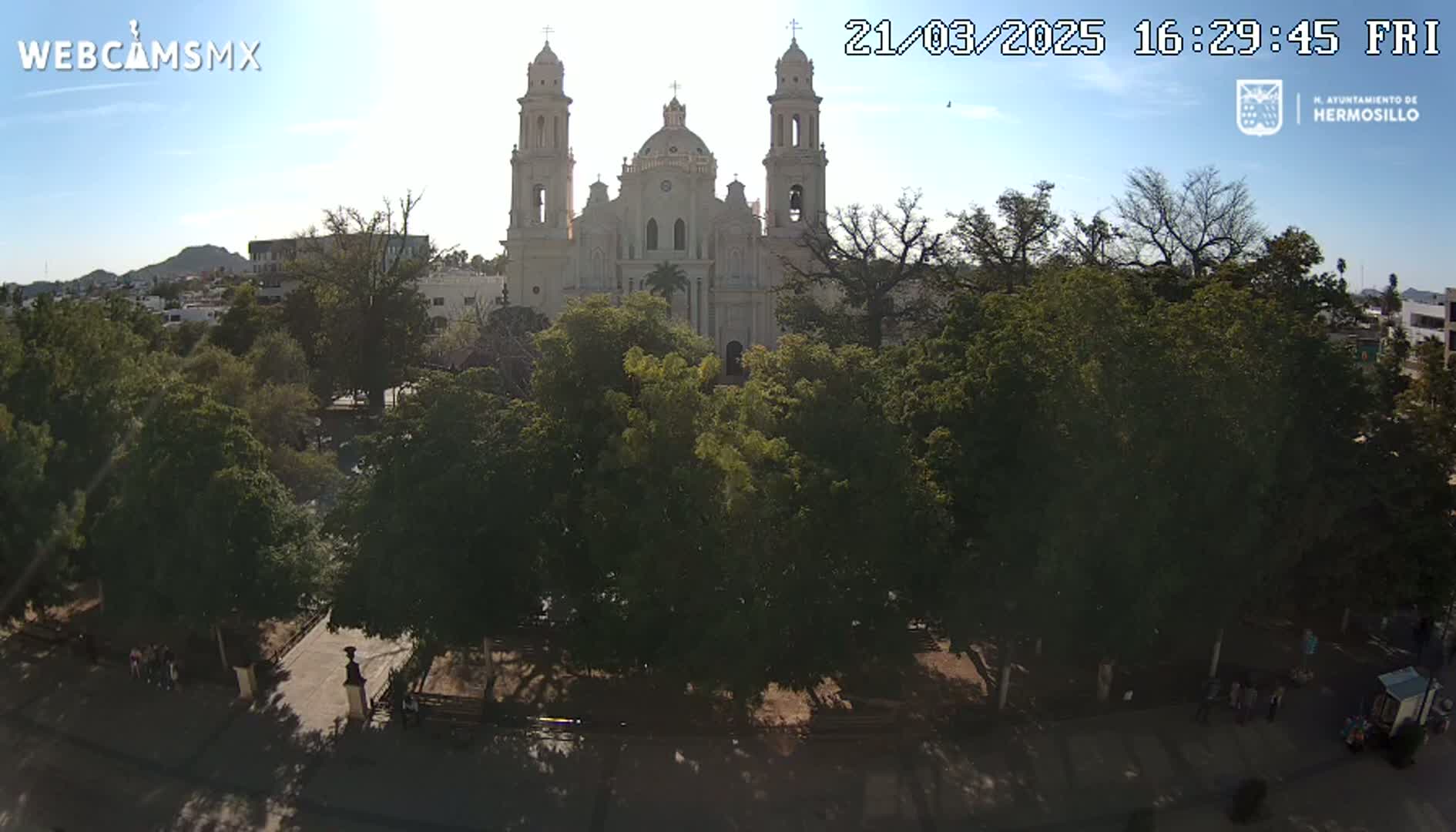 A large cathedral is seen from across a plaza filled with leafy green trees under a mostly sunny sky.