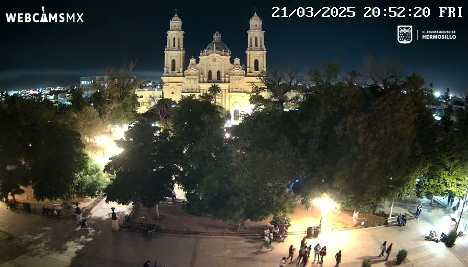 A brightly lit cathedral stands at night in a plaza surrounded by trees and people, under a clear sky.