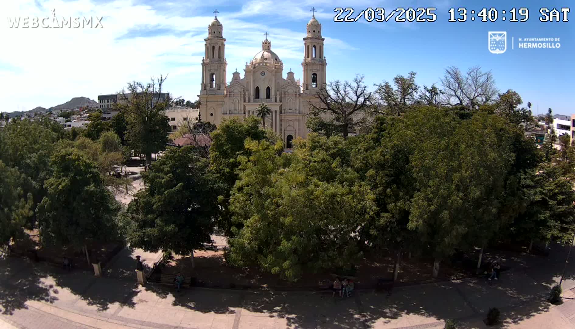A large, light-colored cathedral stands behind a plaza filled with leafy green trees under a mostly sunny sky.