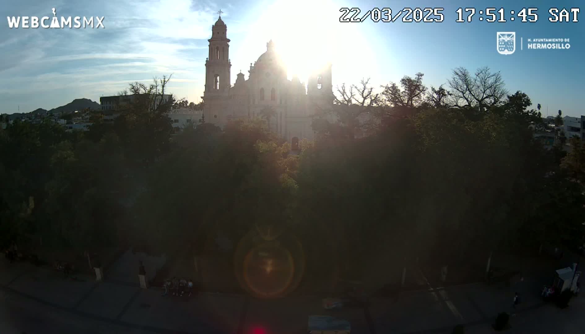 A large, light-colored cathedral is partially obscured by the sun and trees in a city setting at sunset under a clear sky.
