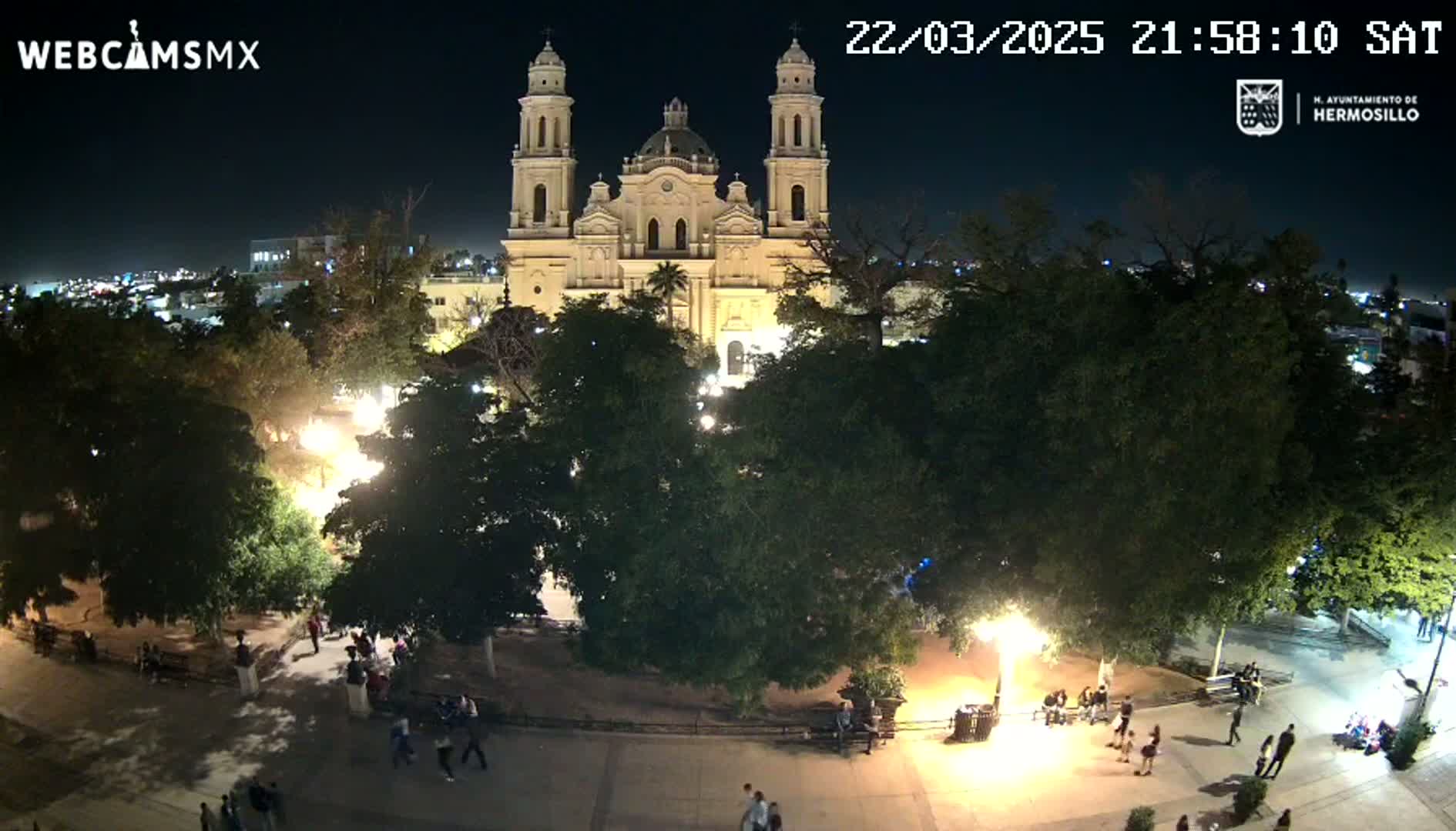 A brightly lit cathedral stands at night behind a plaza with people walking around, under a clear sky.