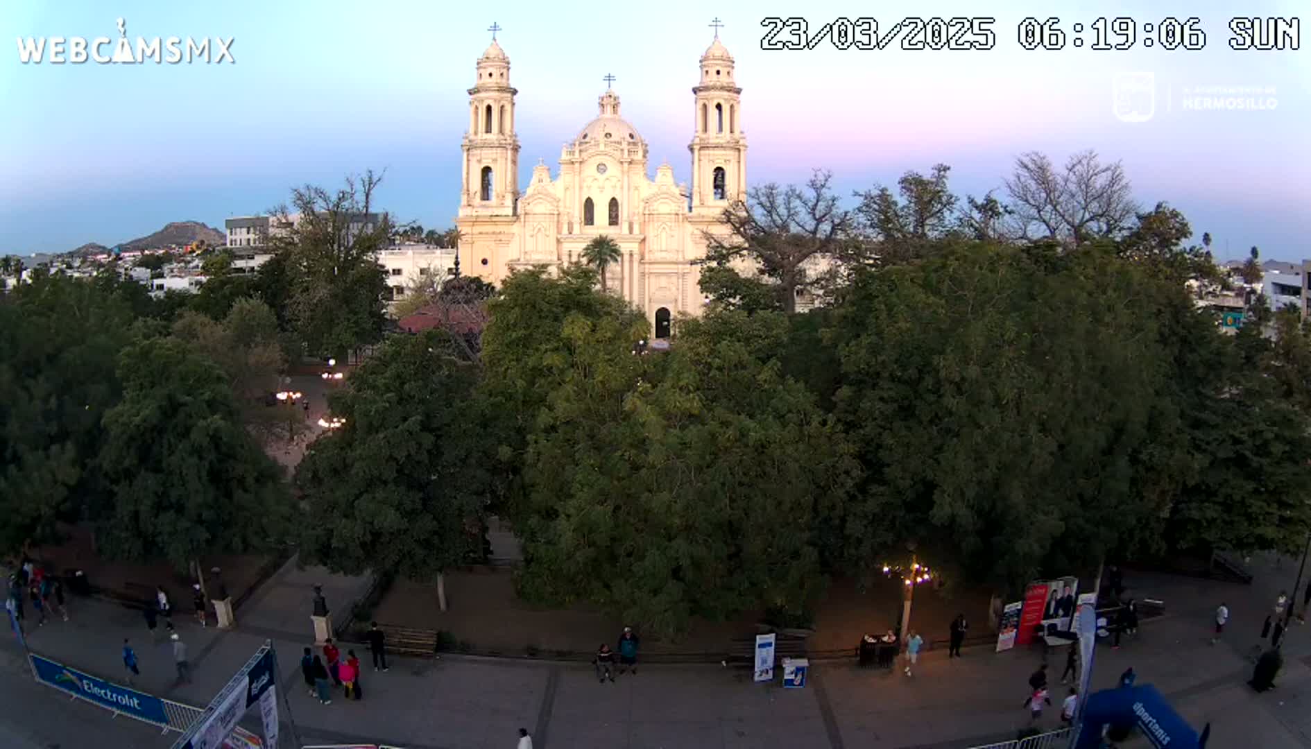A large, light-colored cathedral stands behind a plaza filled with lush green trees under a twilight sky.