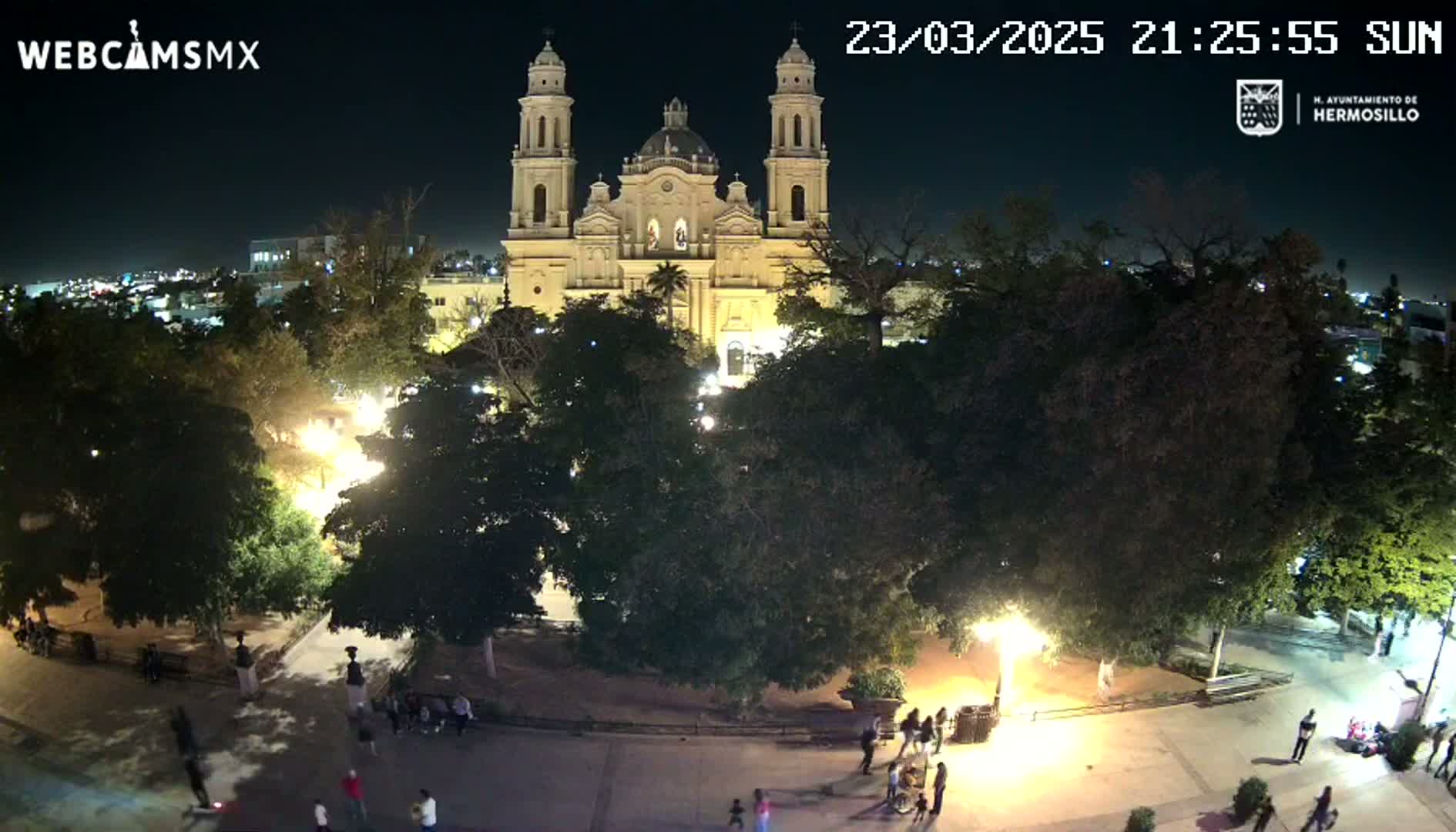 A nighttime, aerial view shows a large plaza with people walking around, surrounded by trees and centered on a brightly lit cathedral.
