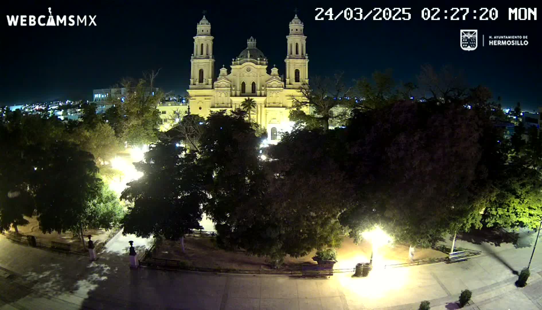 A brightly lit cathedral stands behind a plaza at night, surrounded by dark trees and illuminated walkways.