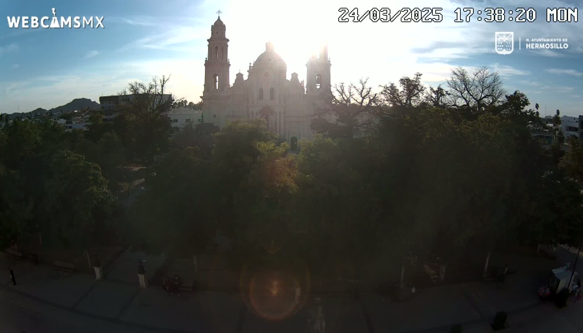 A large, light-colored cathedral stands behind a park filled with lush green trees, all under a partly sunny sky.