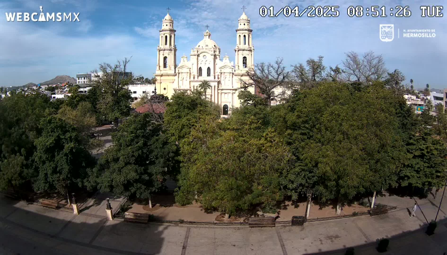 A large cathedral is visible behind a plaza with many leafy green trees and benches under a mostly sunny sky.