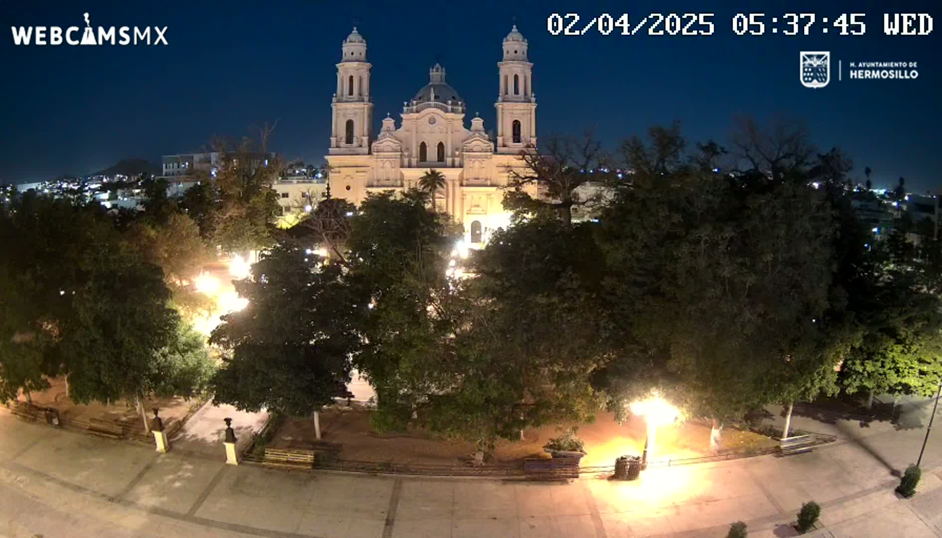 A brightly lit plaza at night, featuring a large cathedral in the background under a clear, dark sky.