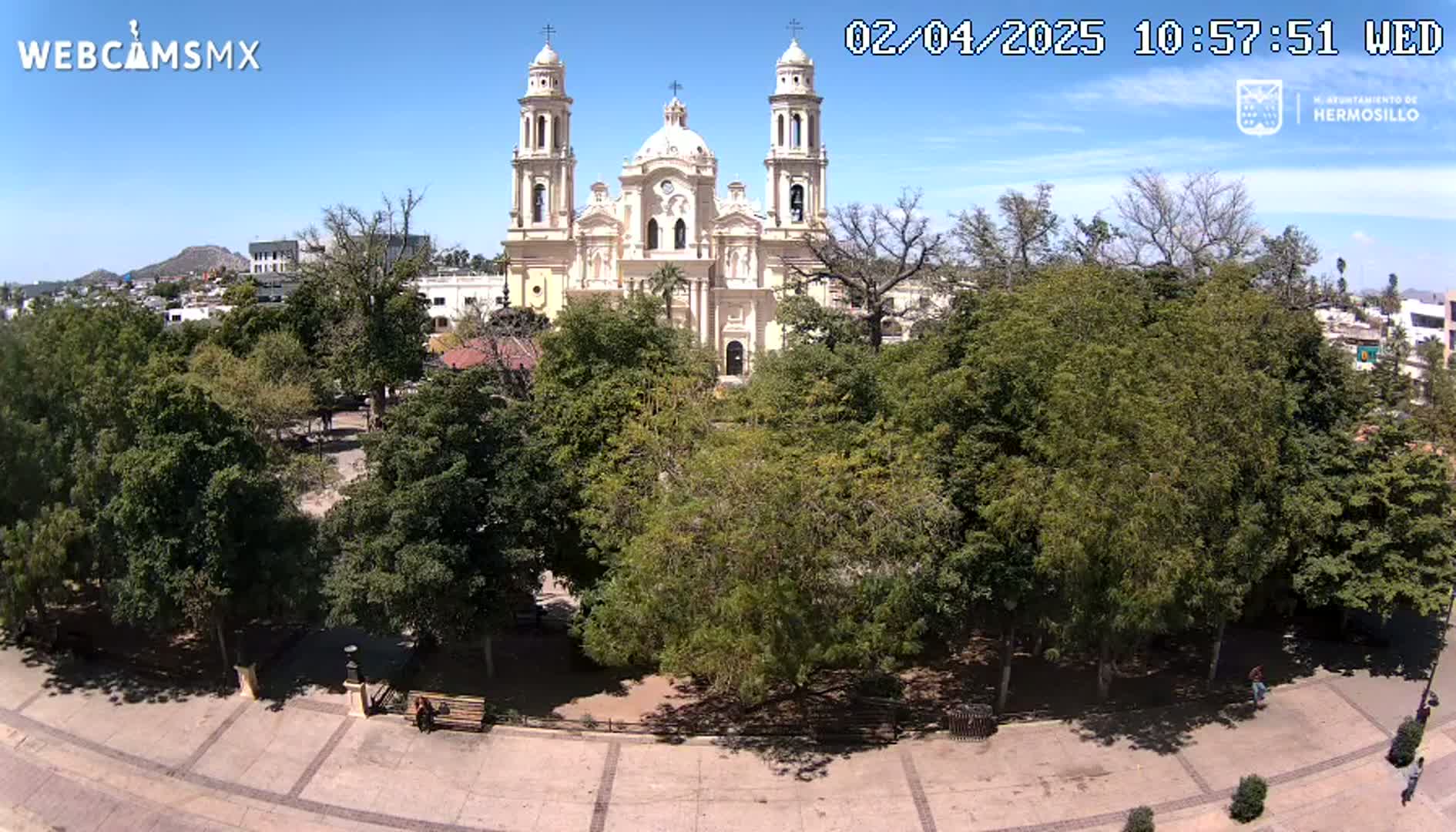A large cathedral stands behind a park filled with lush green trees under a clear blue sky.
