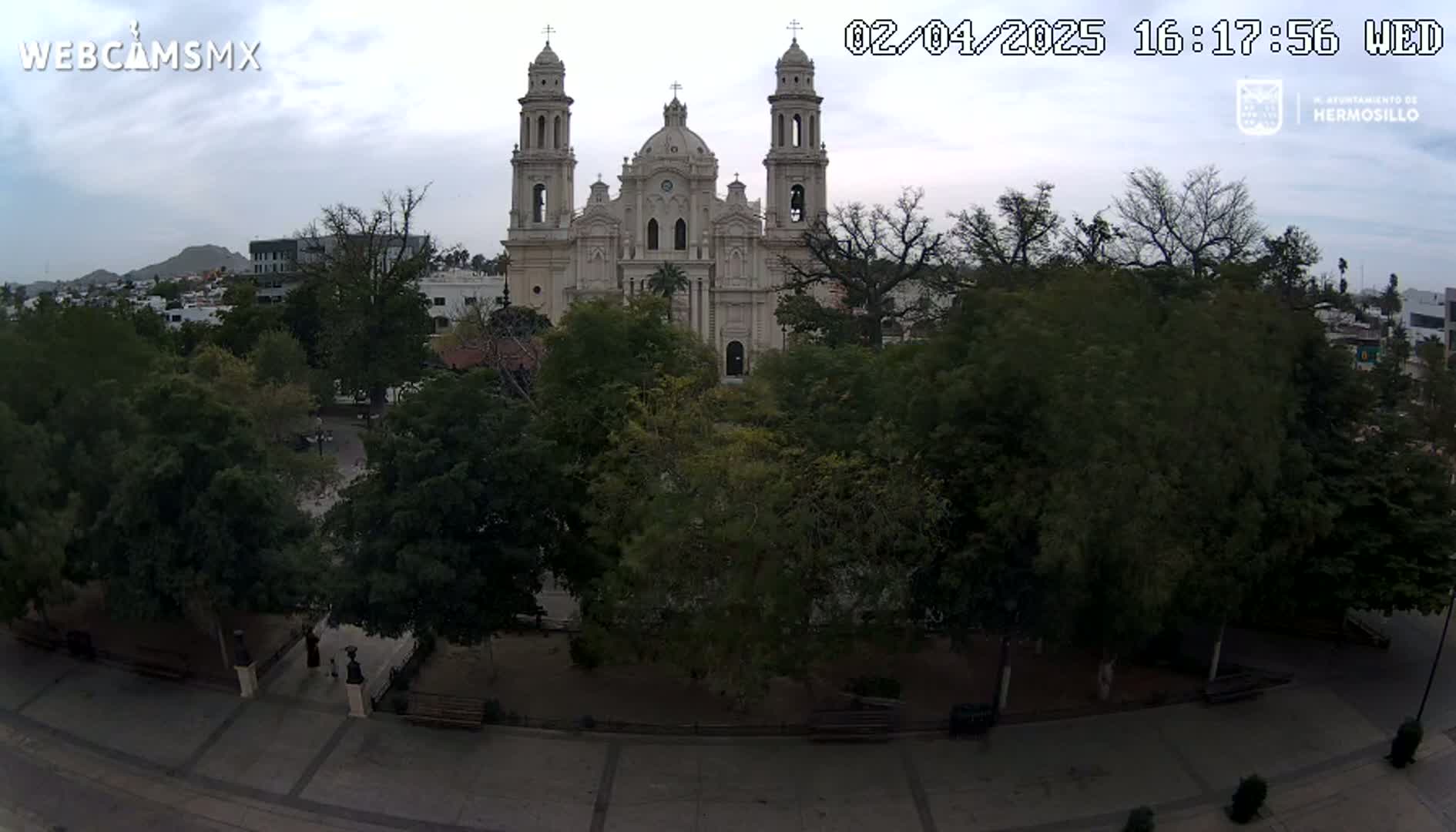 An overcast sky hangs above a large cathedral surrounded by a tree-filled plaza and city buildings.