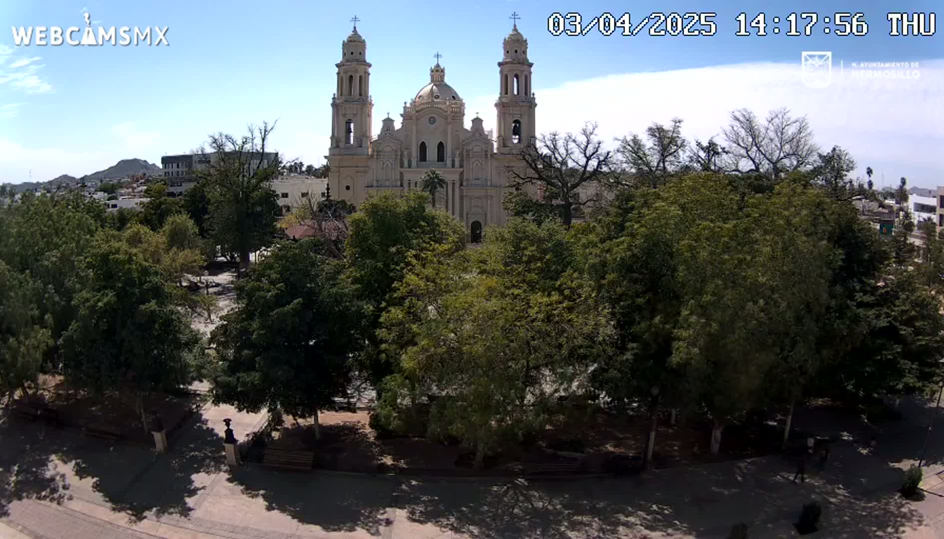 A large cathedral stands behind a tree-filled plaza on a partly sunny day.