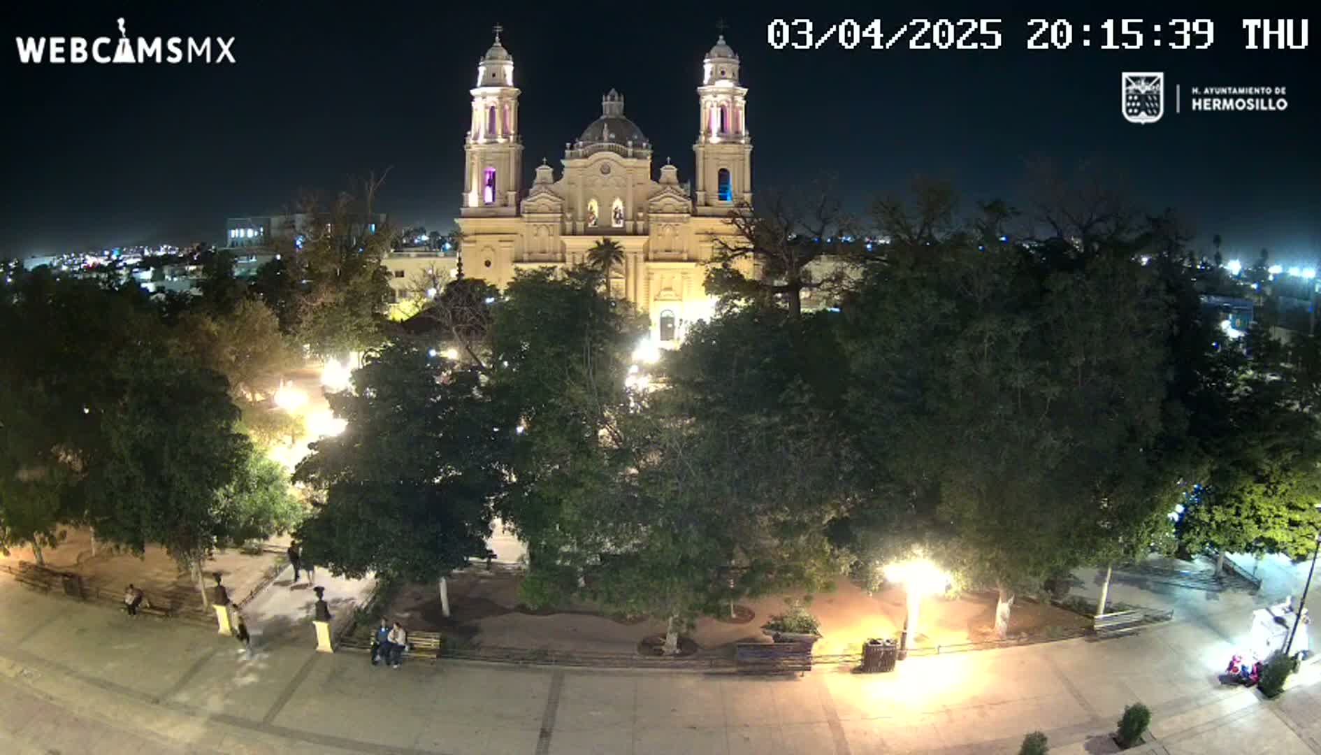 A brightly lit cathedral stands in a nighttime plaza surrounded by trees and a few people; the sky is dark and clear.