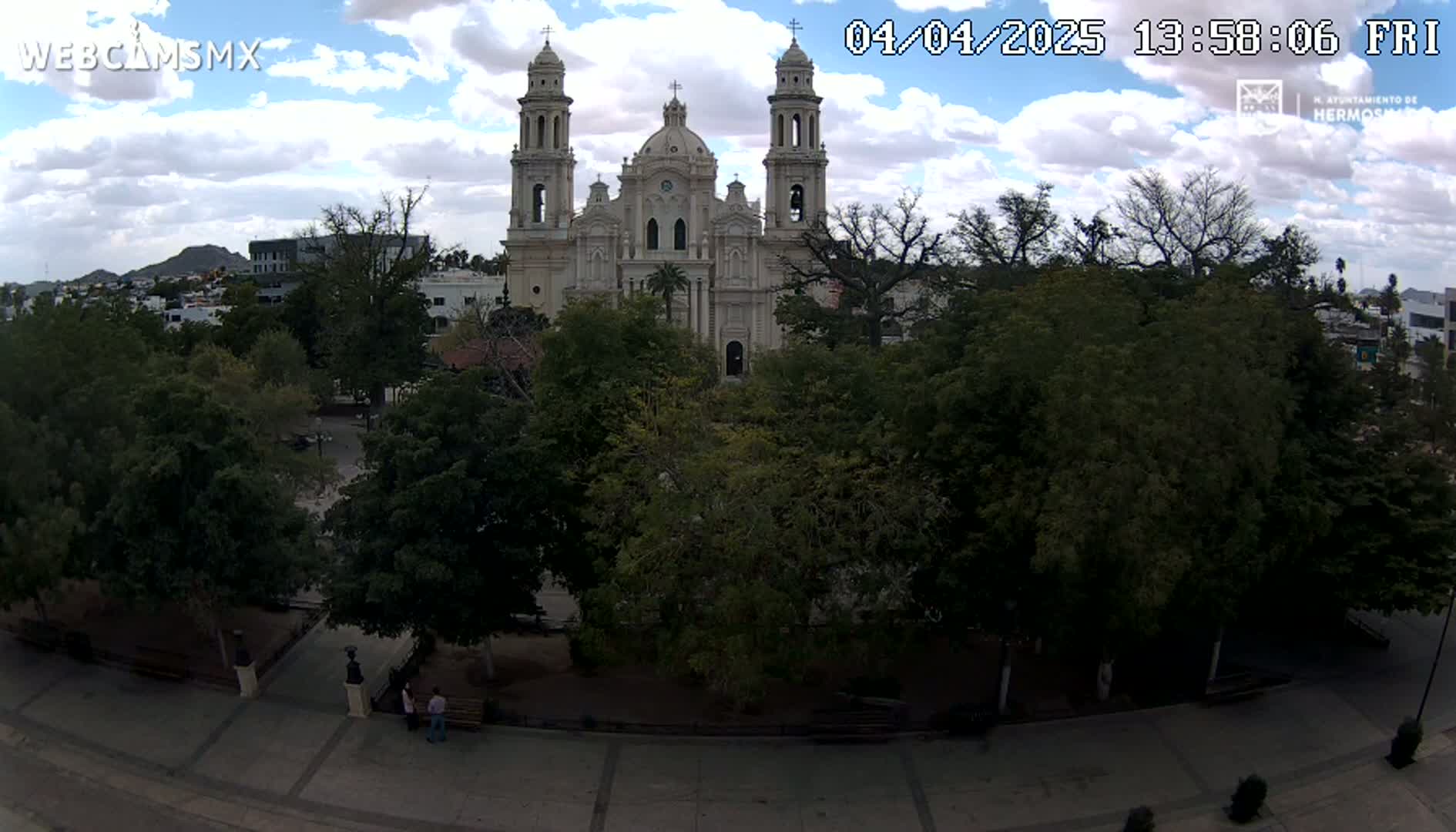 A large, light-colored cathedral is visible behind a plaza filled with lush green trees under a partly cloudy sky.