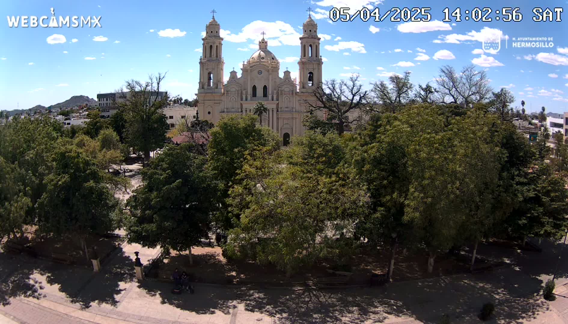 A large cathedral sits behind a plaza filled with leafy green trees under a partly cloudy sky.