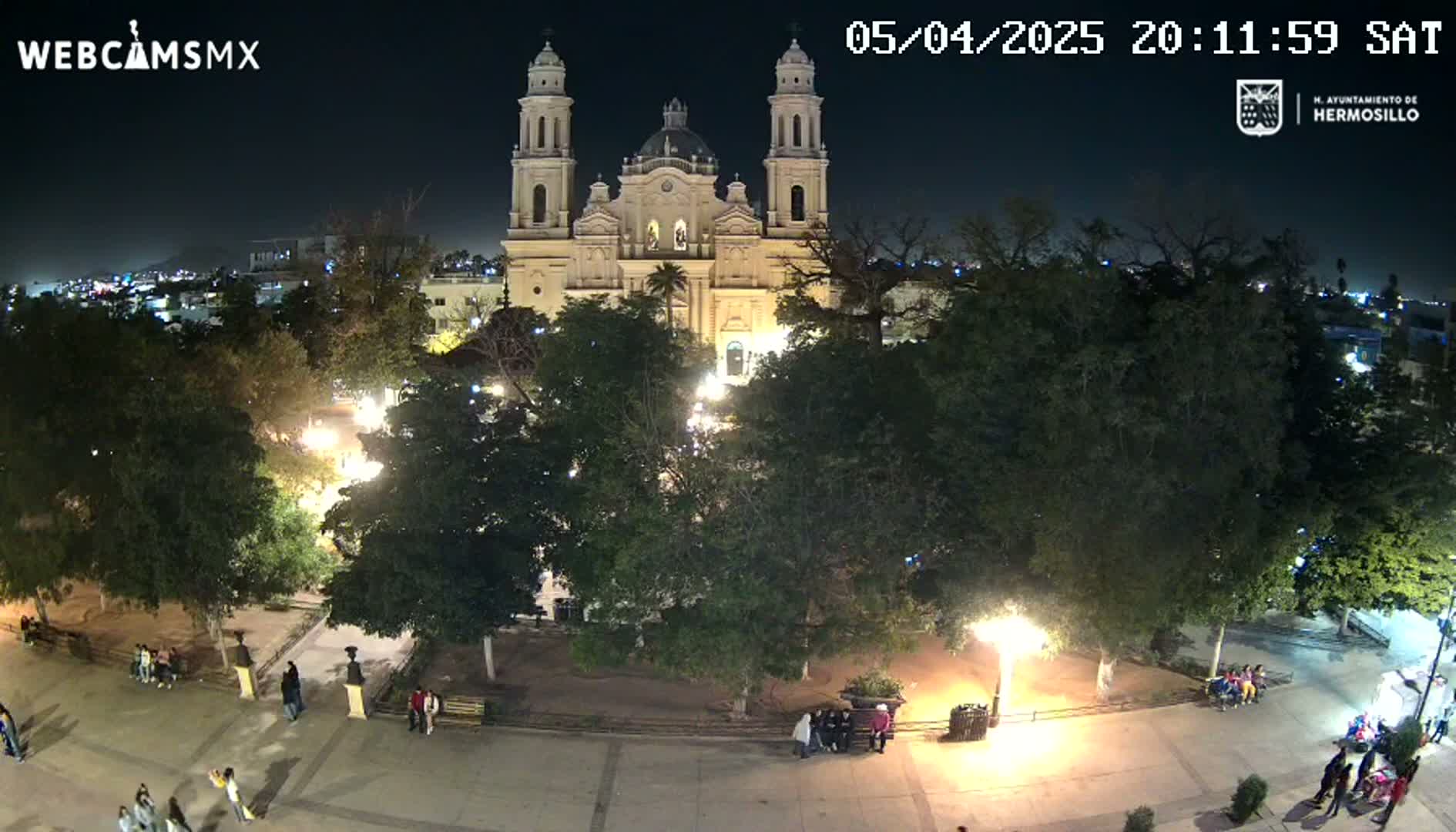 A nighttime aerial view shows a plaza with a large church in the background, surrounded by trees and a few people walking in the square under clear skies.