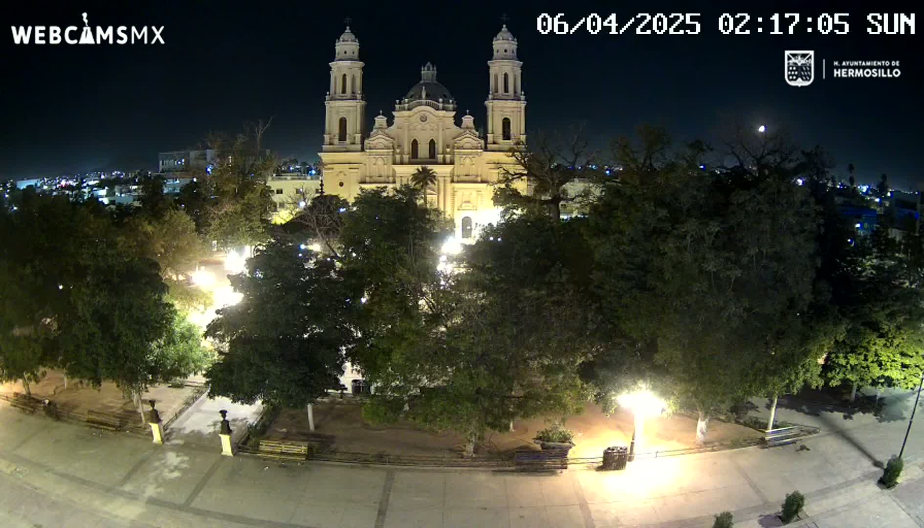 A brightly lit plaza at night, surrounded by trees, with a large cathedral in the background under a clear moonlit sky.