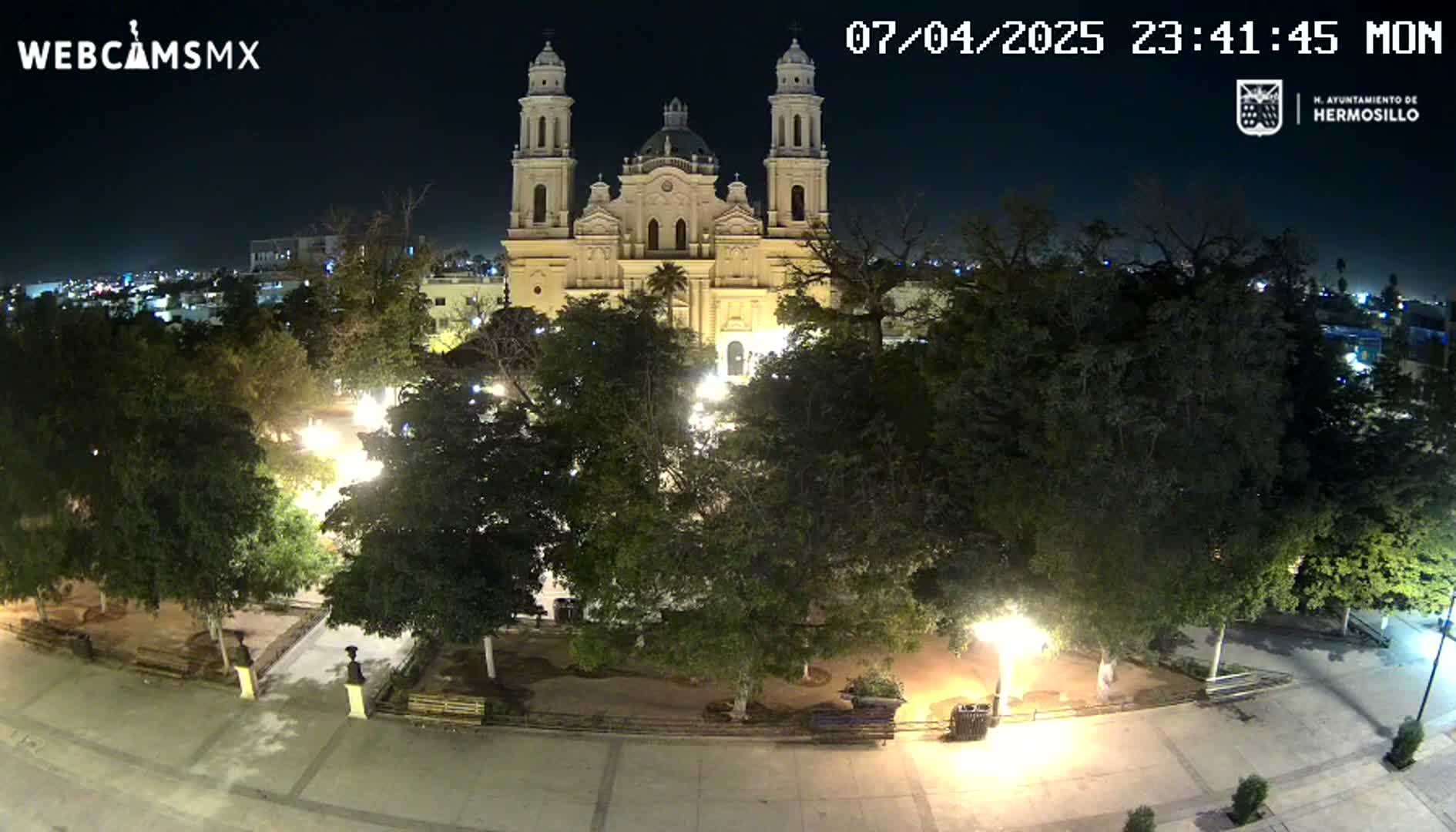 A brightly lit plaza at night, surrounded by trees, features a large cathedral in the background under a clear sky.