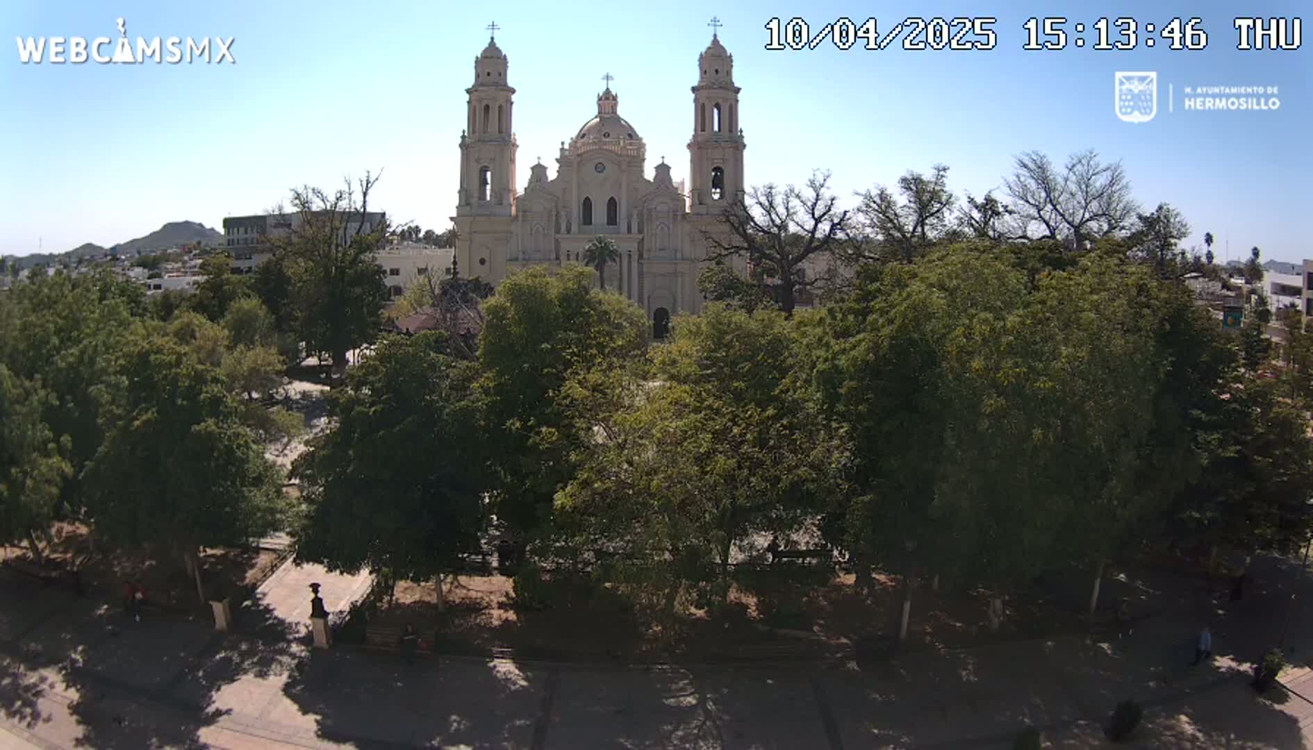 A large cathedral is seen from across a park filled with leafy green trees under a clear, sunny sky.