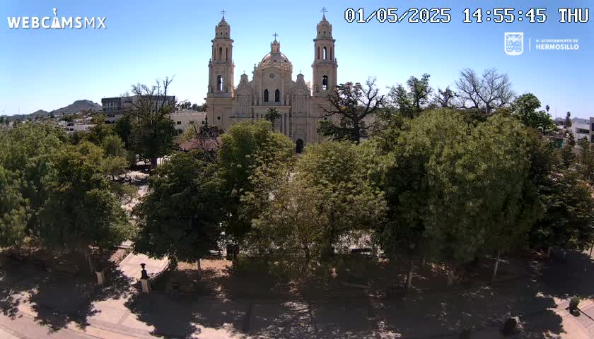 A large cathedral stands in the center of a plaza surrounded by lush green trees under a clear blue sky.
