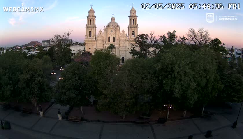 A large, light-colored cathedral stands in a plaza surrounded by lush green trees under a twilight sky.