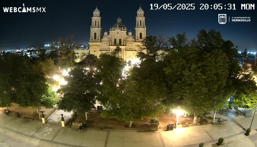 A nighttime, overhead view of a plaza with a large, illuminated cathedral at its center, surrounded by lush green trees and well-lit walkways.