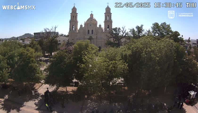 A large cathedral is surrounded by lush green trees in a plaza under a clear blue sky.