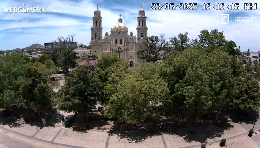 A large cathedral stands in a plaza surrounded by lush green trees under a partly cloudy sky.