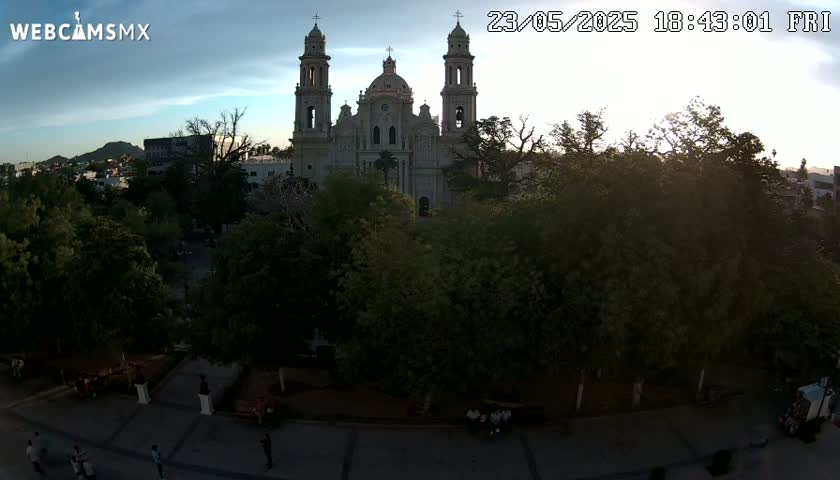 A large cathedral stands in a town square surrounded by lush green trees under a partly cloudy sunset sky.