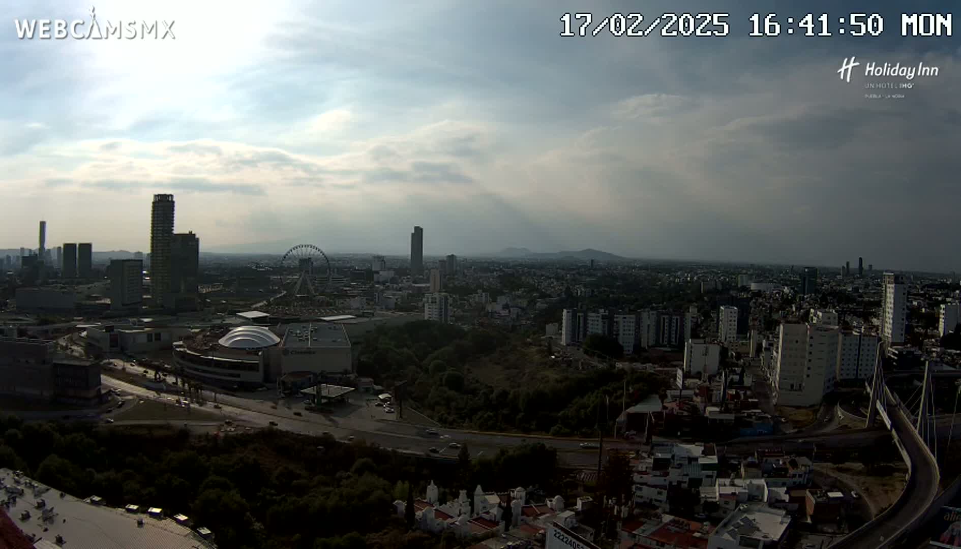 A partly cloudy daytime view of a large city with a Ferris wheel, numerous buildings, and a large highway visible.