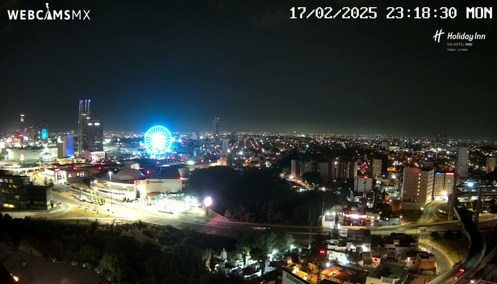 A nighttime aerial view of a city with a brightly lit Ferris wheel and numerous buildings, under a clear sky.