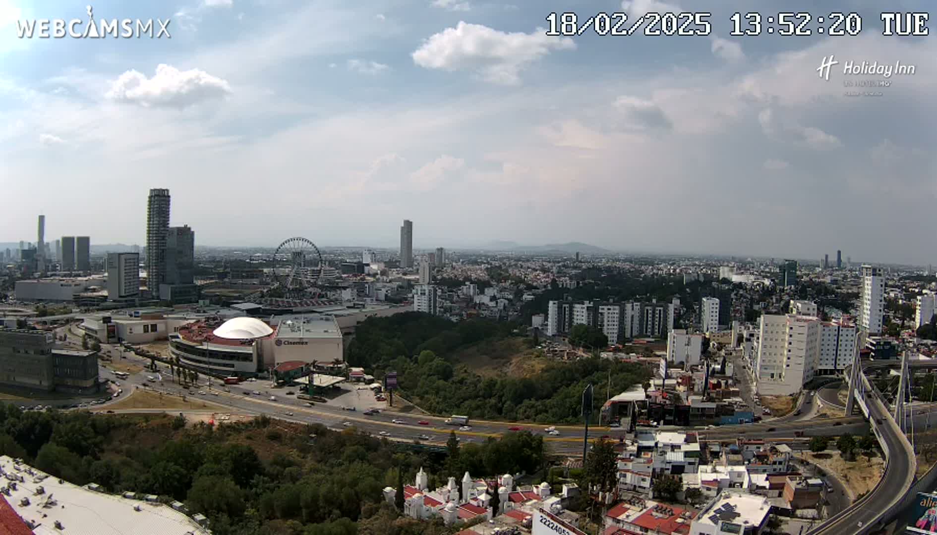 A partly cloudy daytime view of a city with several tall buildings, a Ferris wheel, a large shopping mall, and a highway system.