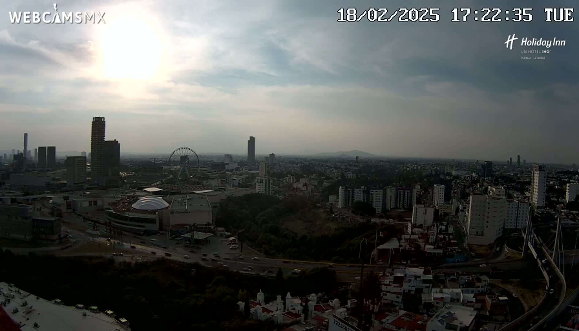 A mostly cloudy, hazy sunset view of a sprawling city with skyscrapers, a Ferris wheel, and a large shopping mall, seen from an elevated vantage point.