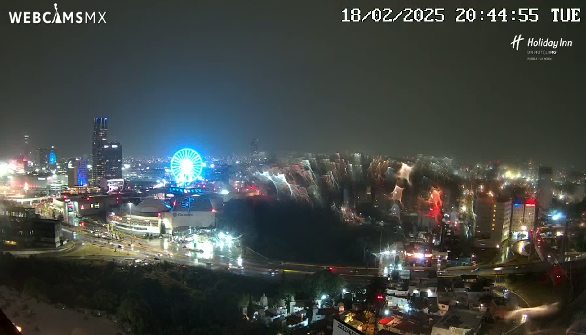 A nighttime, rain-slicked cityscape view shows a Ferris wheel and city lights, with streaks of light possibly from car headlights or other sources in the mid-ground.