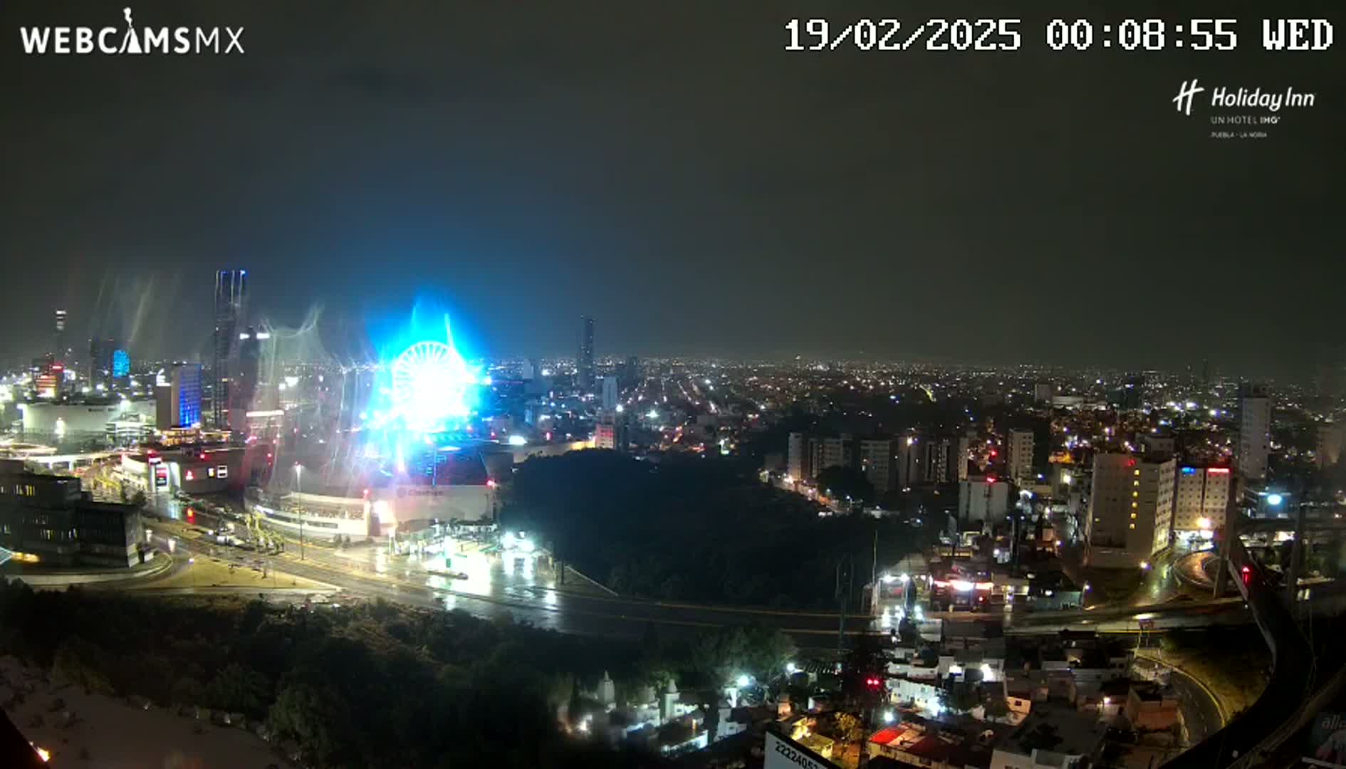 A nighttime, rainy view of a city with a brightly lit Ferris wheel near a large shopping complex.