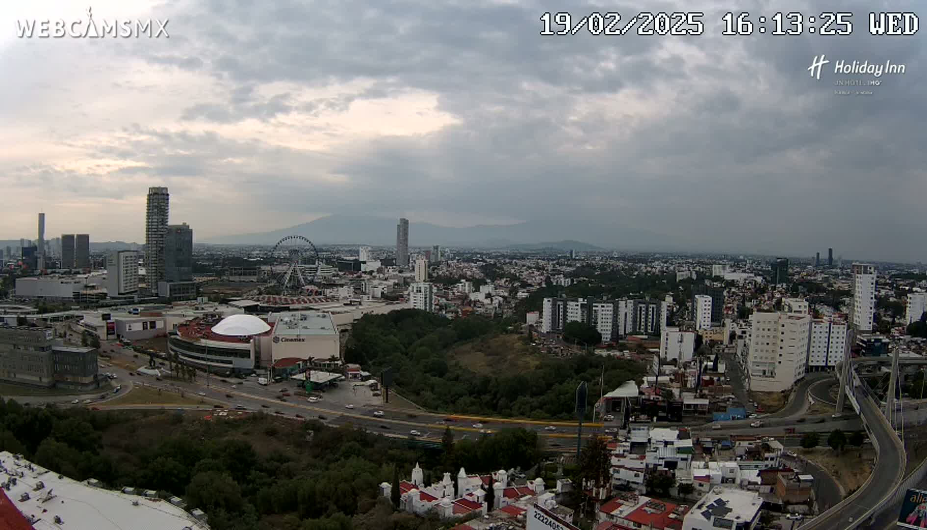 A sprawling city landscape under a cloudy sky, featuring a large Ferris wheel, several high-rise buildings, and a dome-shaped structure near a highway.