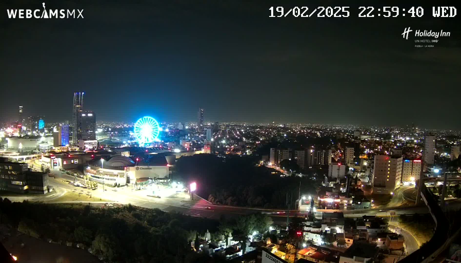 A nighttime, aerial view of a city illuminated by lights, featuring a Ferris wheel and several tall buildings, under a clear sky.