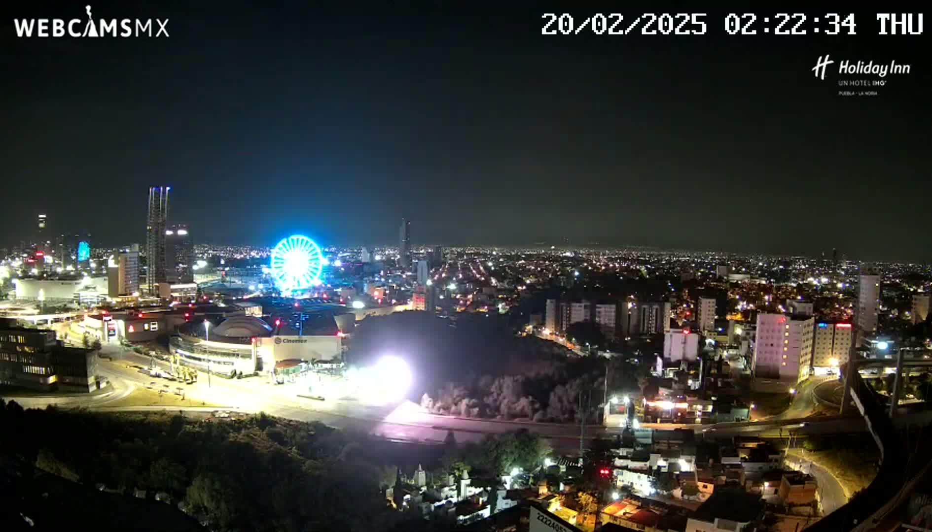 A nighttime view of a city illuminated with lights, featuring a Ferris wheel and a demolition or construction event in progress.