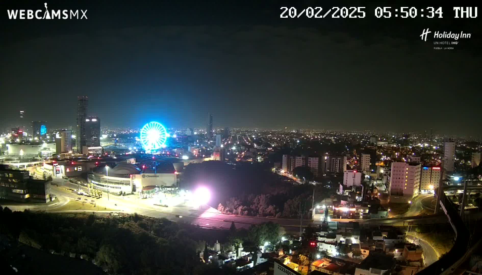 A nighttime aerial view of a city with a brightly lit Ferris wheel and surrounding buildings, under a clear sky.
