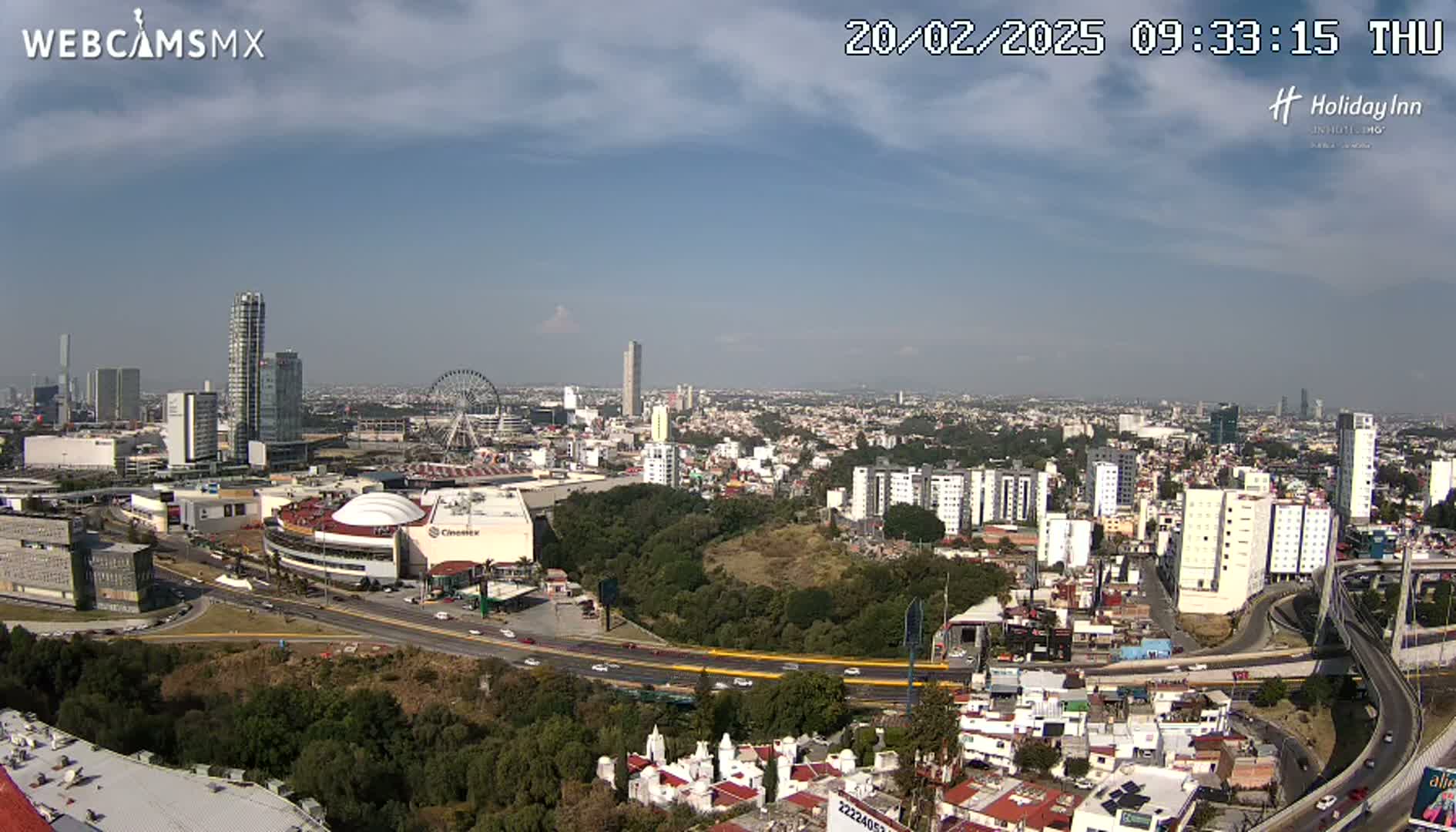 A partly cloudy daytime view of a sprawling city, featuring a Ferris wheel, several tall buildings, and a large highway system.