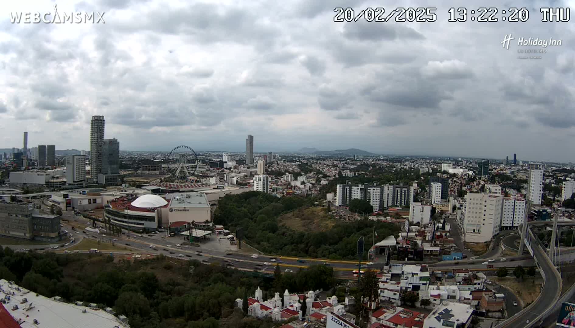 A sprawling city scene under a cloudy sky shows a large shopping mall, a Ferris wheel, many high-rise buildings, and a highway system.