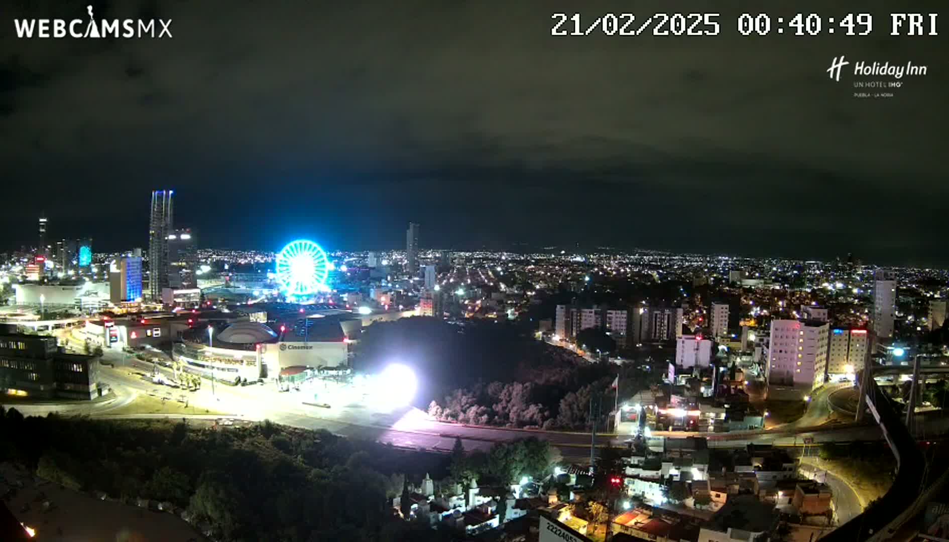 A nighttime aerial view of a city with a brightly lit Ferris wheel and several tall buildings, under a mostly clear sky.