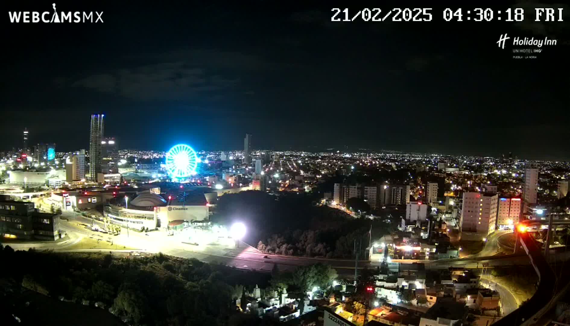 A nighttime aerial view of a city illuminated by lights, featuring a Ferris wheel and various buildings, under a clear, dark sky.